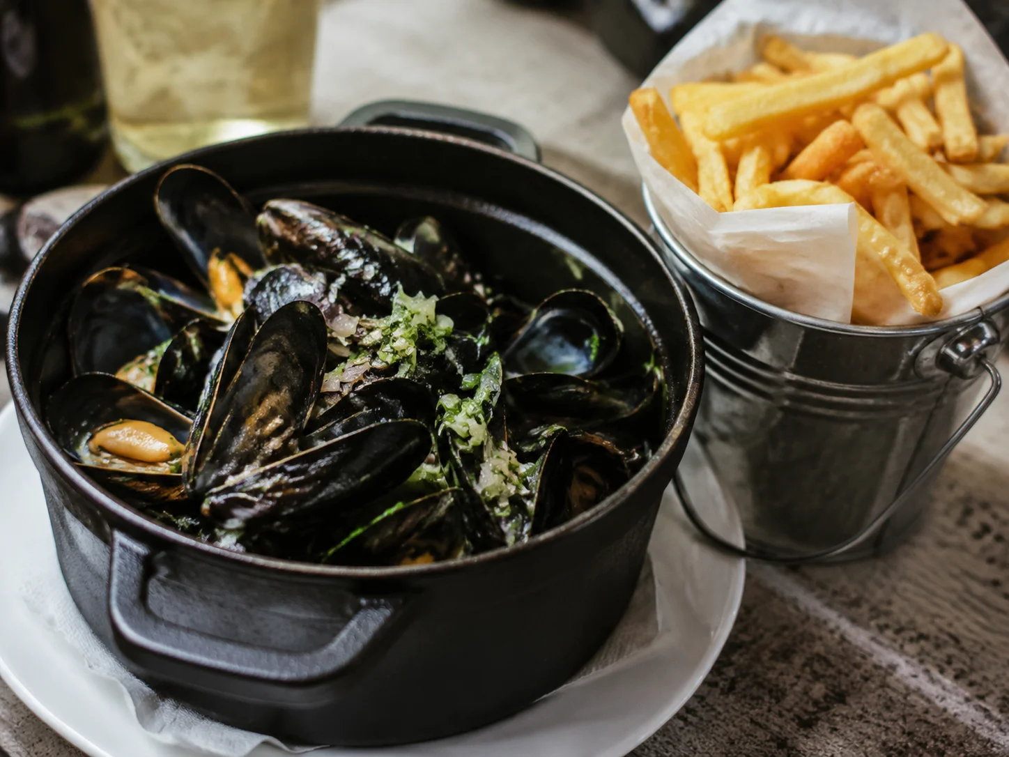 Close-up-Aufnahme eines schwarzen Gusseisen-Topfs mit Muscheln (Moules mariniere) und einer kleinen Schale goldener belgischer Frites, professionelle Food-Photography